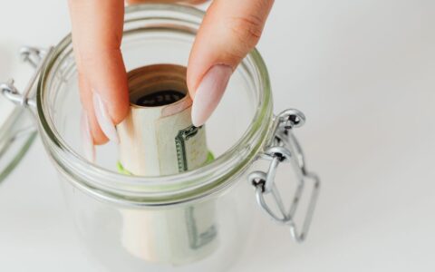 Close-up of hand placing rolled currency into a clear glass jar, symbolizing savings.