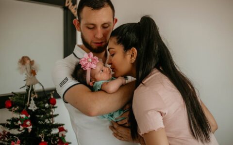 A loving couple kisses their baby beside a decorated Christmas tree.