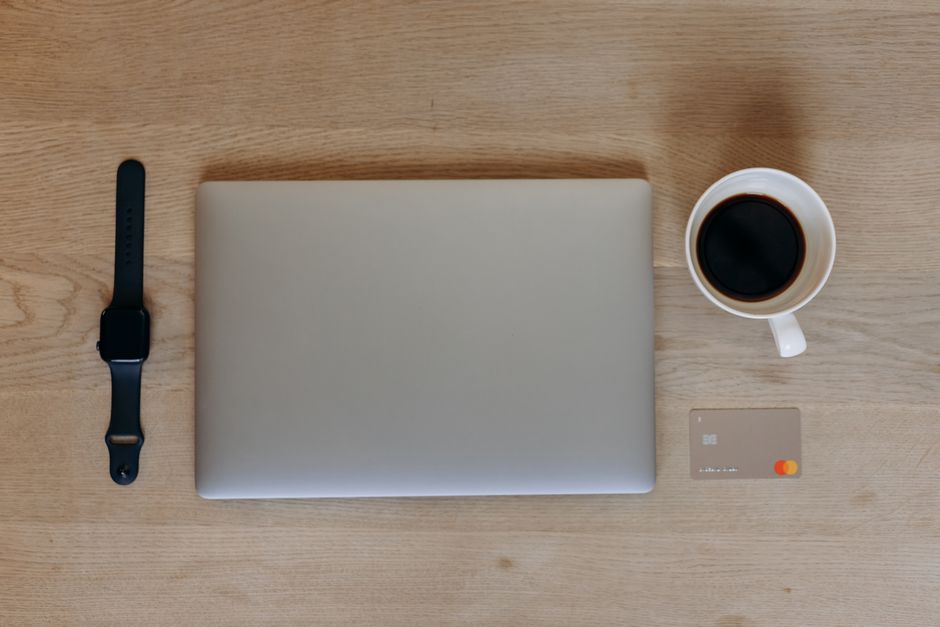 Overhead view of a sleek desk with a laptop, smartwatch, coffee, and credit card on a wooden surface.
