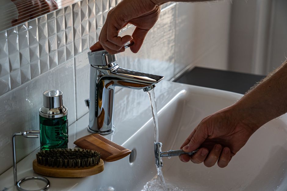 A man's hands adjust the faucet in a bathroom, surrounded by grooming tools like a comb and brush.