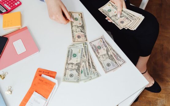 A person counting US dollar bills at a white desk with office supplies nearby.