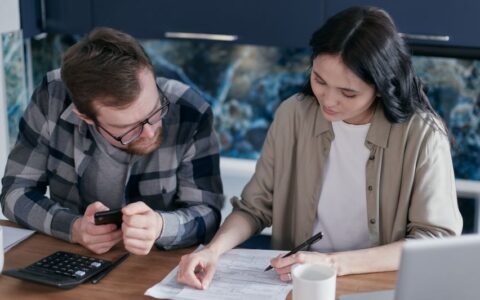 A couple reviewing financial documents at a table, using gadgets like a calculator and phone.