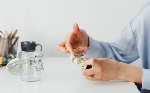 Close-up of hands rolling US dollars with a rubber band near an empty glass jar.