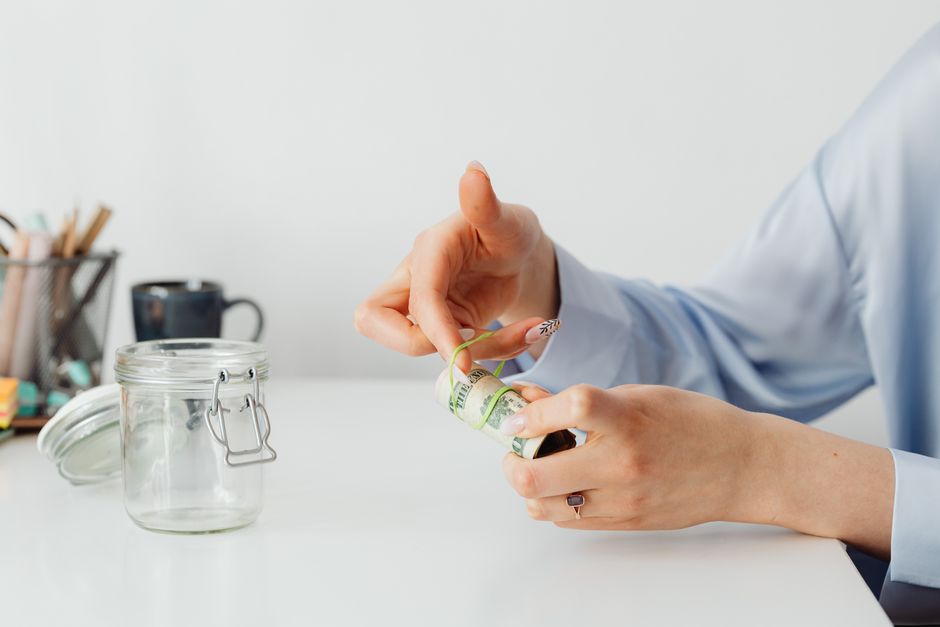 Close-up of hands rolling US dollars with a rubber band near an empty glass jar.