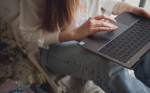 Crop unrecognizable female in casual clothes sitting on couch and using touchpad on laptop at home