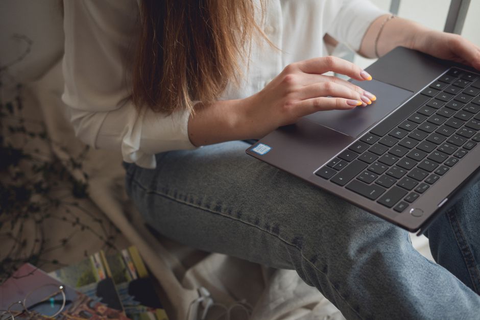Crop unrecognizable female in casual clothes sitting on couch and using touchpad on laptop at home