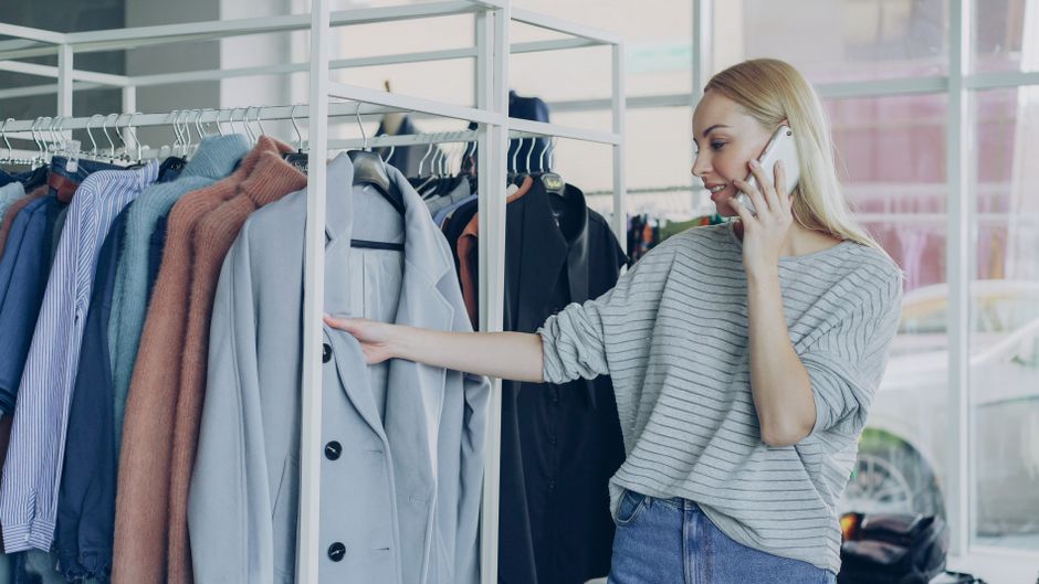 Young woman shopping for clothes while talking on her mobile phone. Bright indoor fashion scene.
