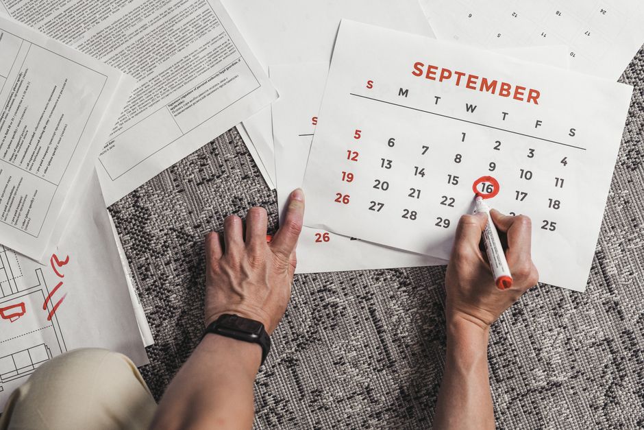 Hands holding a calendar with September dates encircled, surrounded by documents on a carpet.