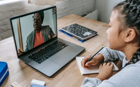 From above of focused Asian girl sitting at table with copybook and pencil and carefully listening to black teacher explaining homework task during videocall on laptop