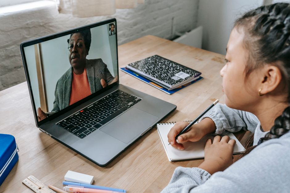 From above of focused Asian girl sitting at table with copybook and pencil and carefully listening to black teacher explaining homework task during videocall on laptop