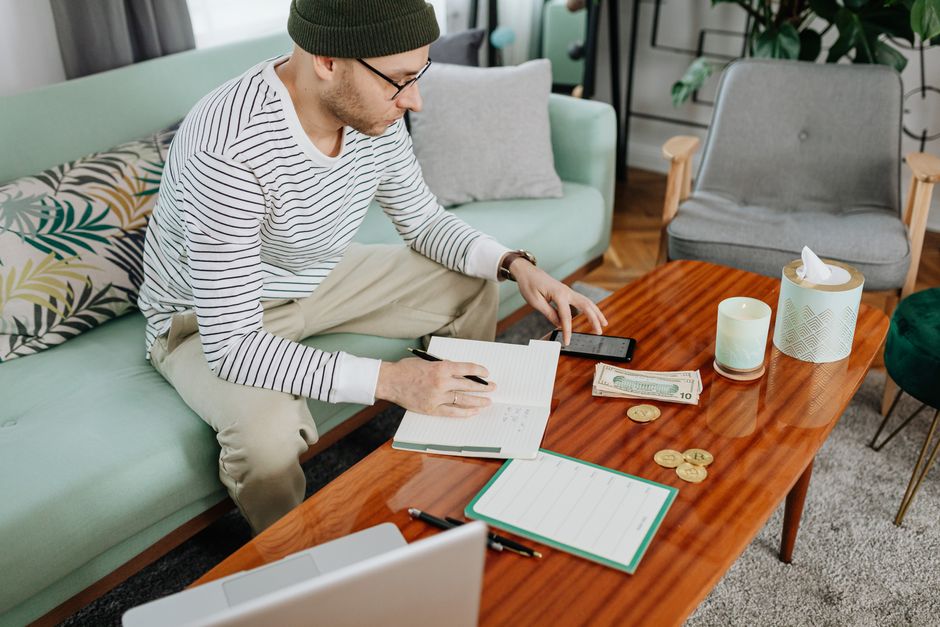 A man in casual attire managing finances with a laptop and notebook on a wooden table indoors.